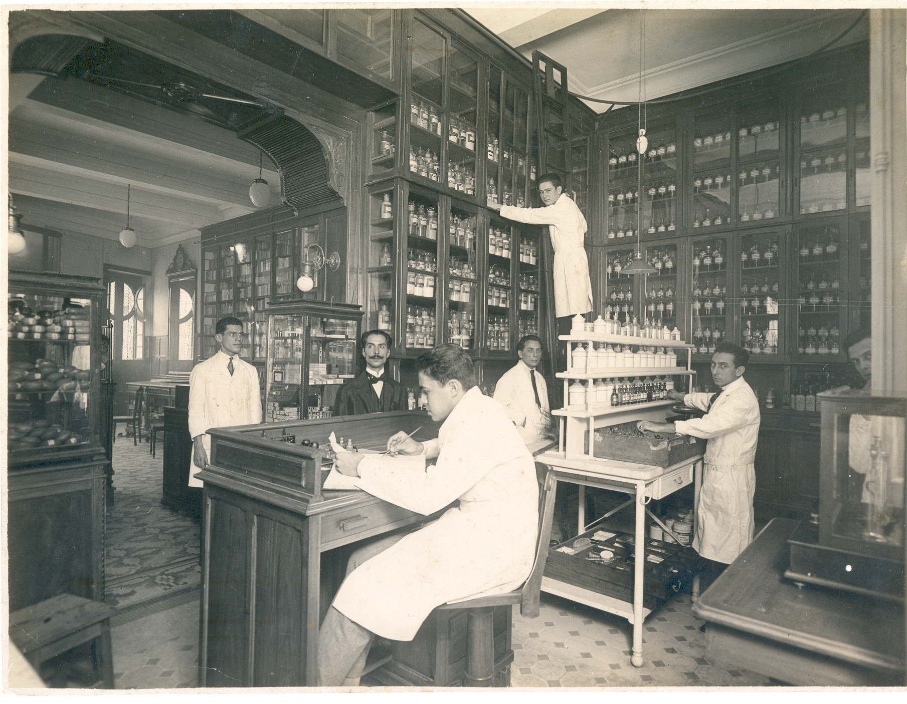 Foto em preto e branco do interior de uma farmácia de 1910 com armários com portas de vidro que vão do chão até o teto. No centro, há uma estação de manipulação de remédios e uma escrivaninha. Há seis homens no interior vestidos com jalecos e um deles está no alto de uma escada encostada no armário, e outro homem está vestido com terno e gravata borboleta. 