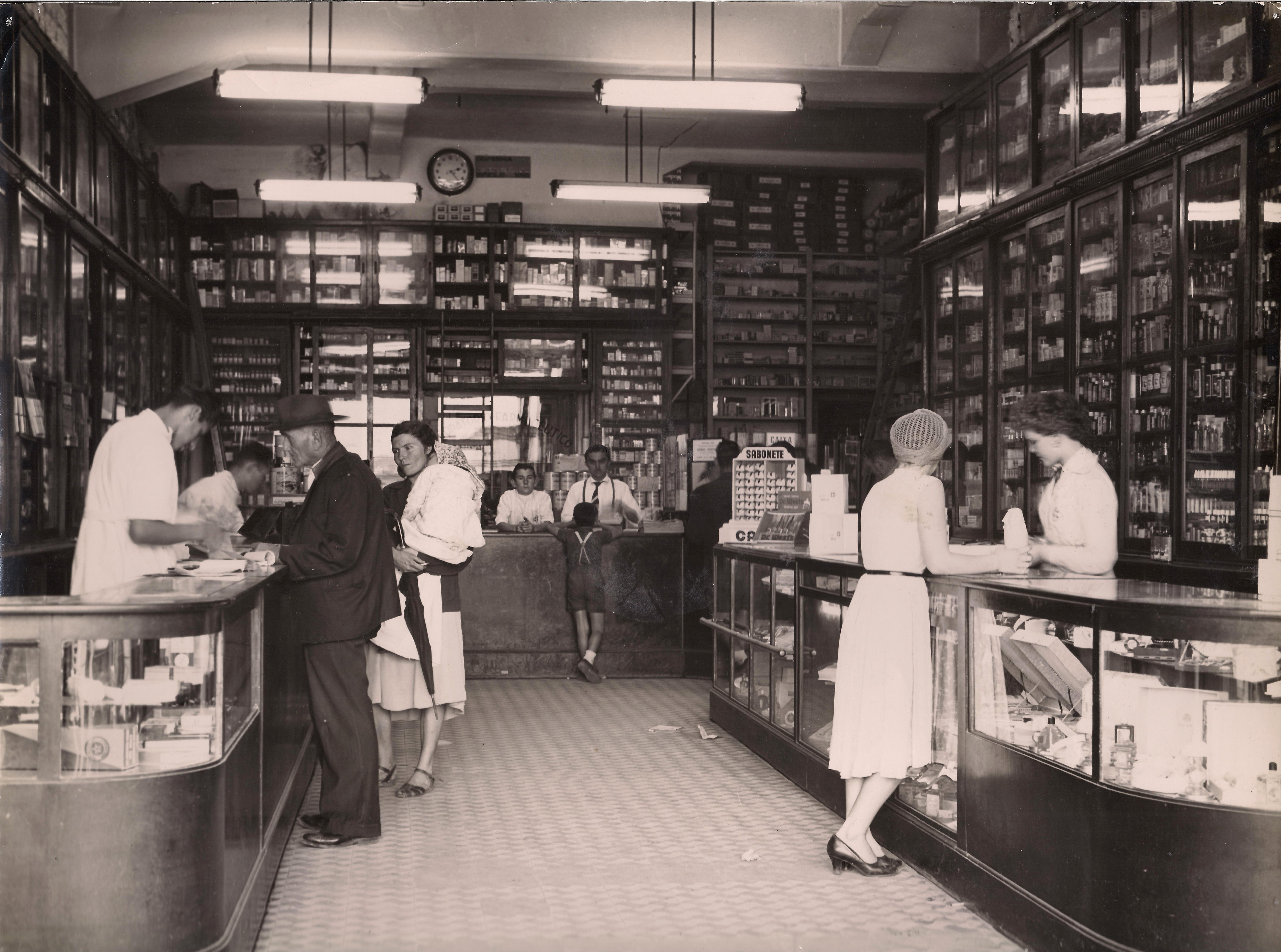 Foto em preto e branco do interior de uma farmácia de 1950 com armários com portas de vidro que vão do chão até o teto. Há três balcões de atendimento com parte da sua estrutura em vidro para mostrar mais medicamentos. Há homens e mulheres atrás e na frente dos balções comprando e vendendo produtos.