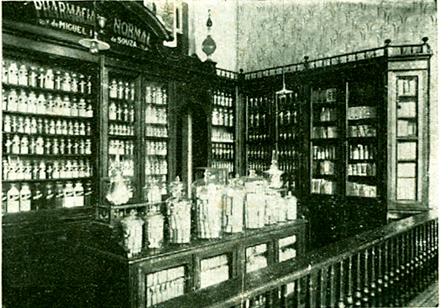Foto antiga e em preto e branco do interior da Pharmacia Normal Drogaria e Perfumaria de Piracicaba/SP. Há diversas prateleiras com frascos de vidros sobre elas.