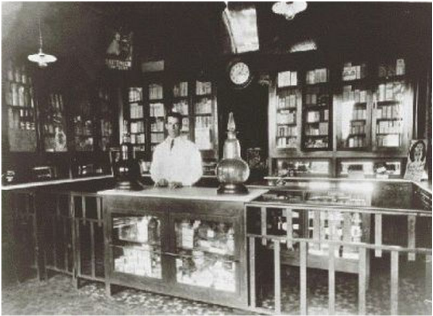Foto antiga e em preto e branco do interior da farmácia Santa Terezinha. Há um homem de jaleco atrás do balcão e diversas prateleiras e balcões dentro dela.