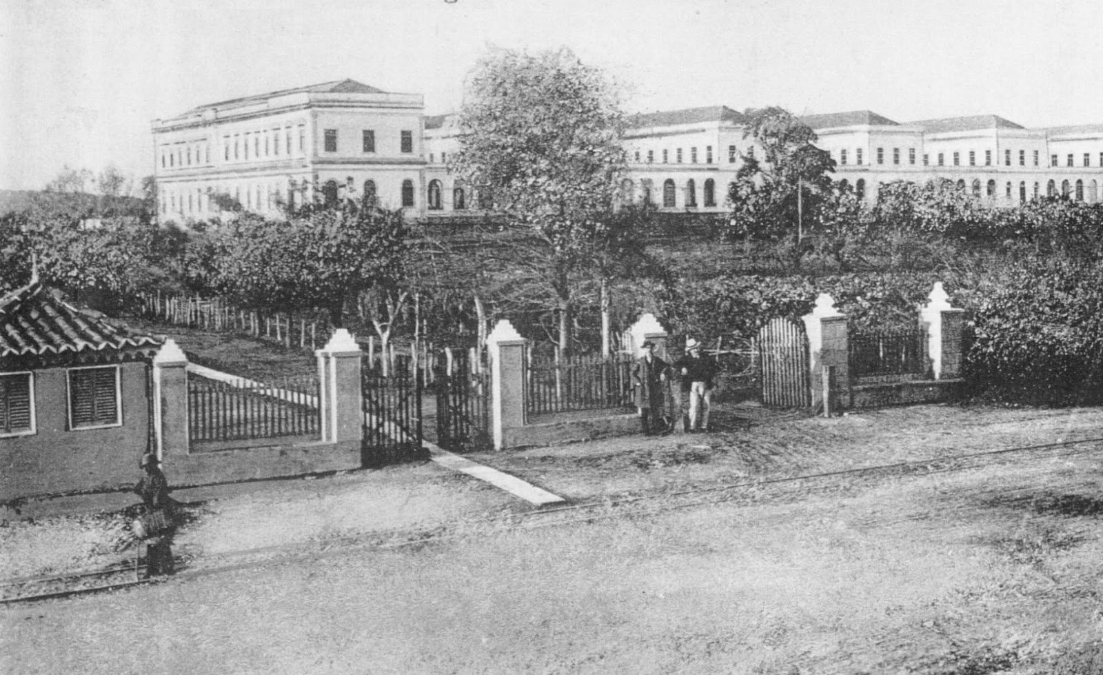 Foto antiga e em preto e branco da entrada do hospício São Pedro, de Porto Alegre, no Rio Grande do Sul. Há uma grade com portões no meio, uma vasta área com arvores e, ao fundo, diversos sobrados lado a lado. 
