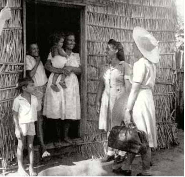 Foto antiga e em preto e branco de uma mulher com crianças na porta de uma casa simples de sapê. Do lado de fora, estão outras duas mulheres conversando com eles. Uma delas carrega uma maleta em suas mãos. 