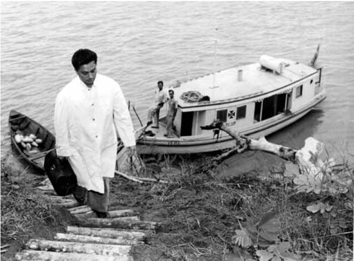Foto antiga e em preto e branco de um home de jaleco branco subindo as escadas da margem de um rio. Na água, há um pequeno barco ancorado com dois homens dentro dele.
