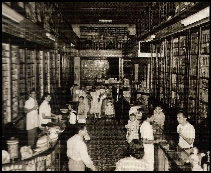 Foto antiga e em preto e branco de diversas pessoas dentro da Farmácia Oswaldo Cruz. É um ambiente amplo com armários cheios de prateleiras, que tomam o fundo e as paredes laterais. 