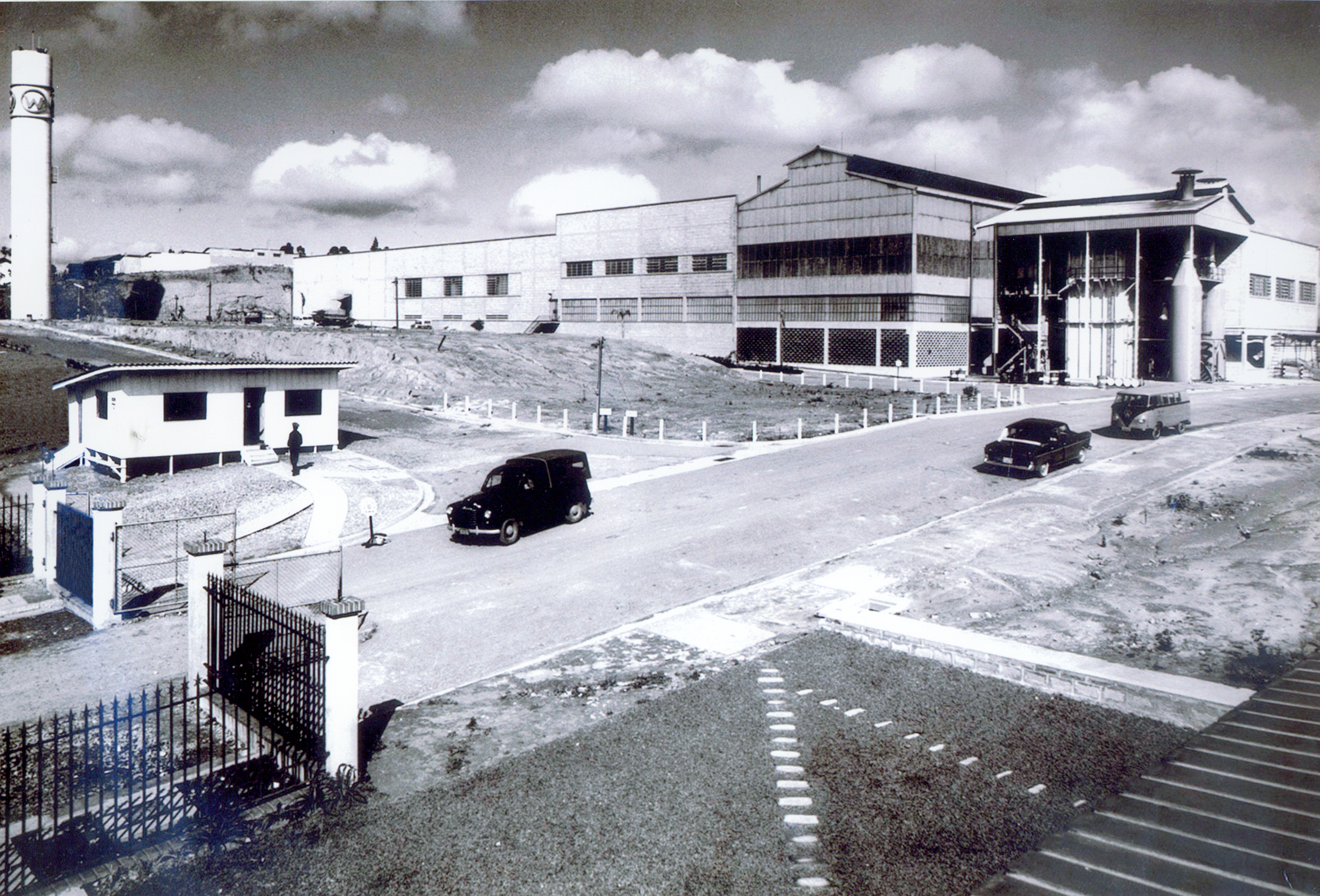 Foto antiga e em preto e branco de um terreno com diversas construções. Há pessoas e carros passando. 