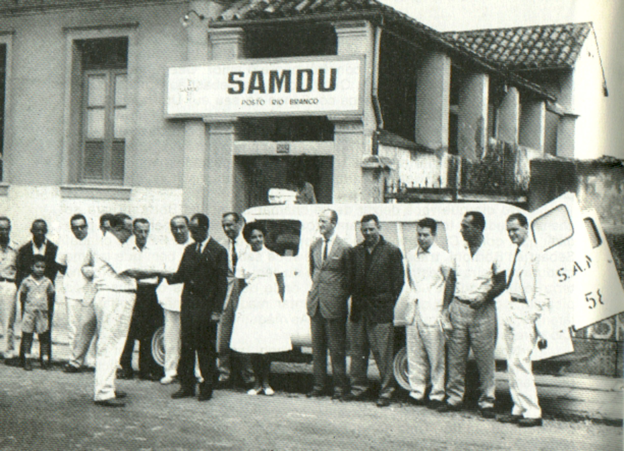 Foto em preto e branco de um grupo de pessoas em frente a uma ambulância com o Hospital SAMDU ao fundo.