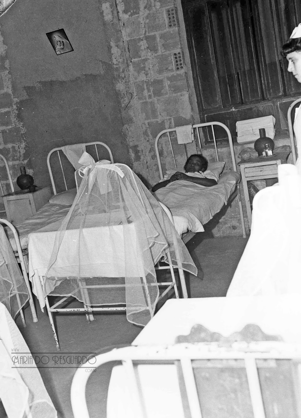 Foto em preto e branco do interior de um quarto de hospital com quatro camas enfileiradas, e em uma delas há uma mulher deitada.