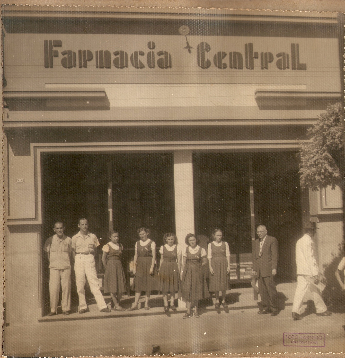 Foto antiga da fachada da Farmácia Central em Crato, Ceará. Ela está com a porta aberta, e na sua frente há um grupo de homens e meninas, todos em pé.