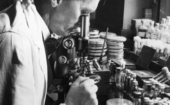 Foto do cientista Alexander Fleming dentro de um laboratório. Ele olha pela lente de um microscópio e ao seu redor há vários instrumentos, tubos de ensaio e placas.