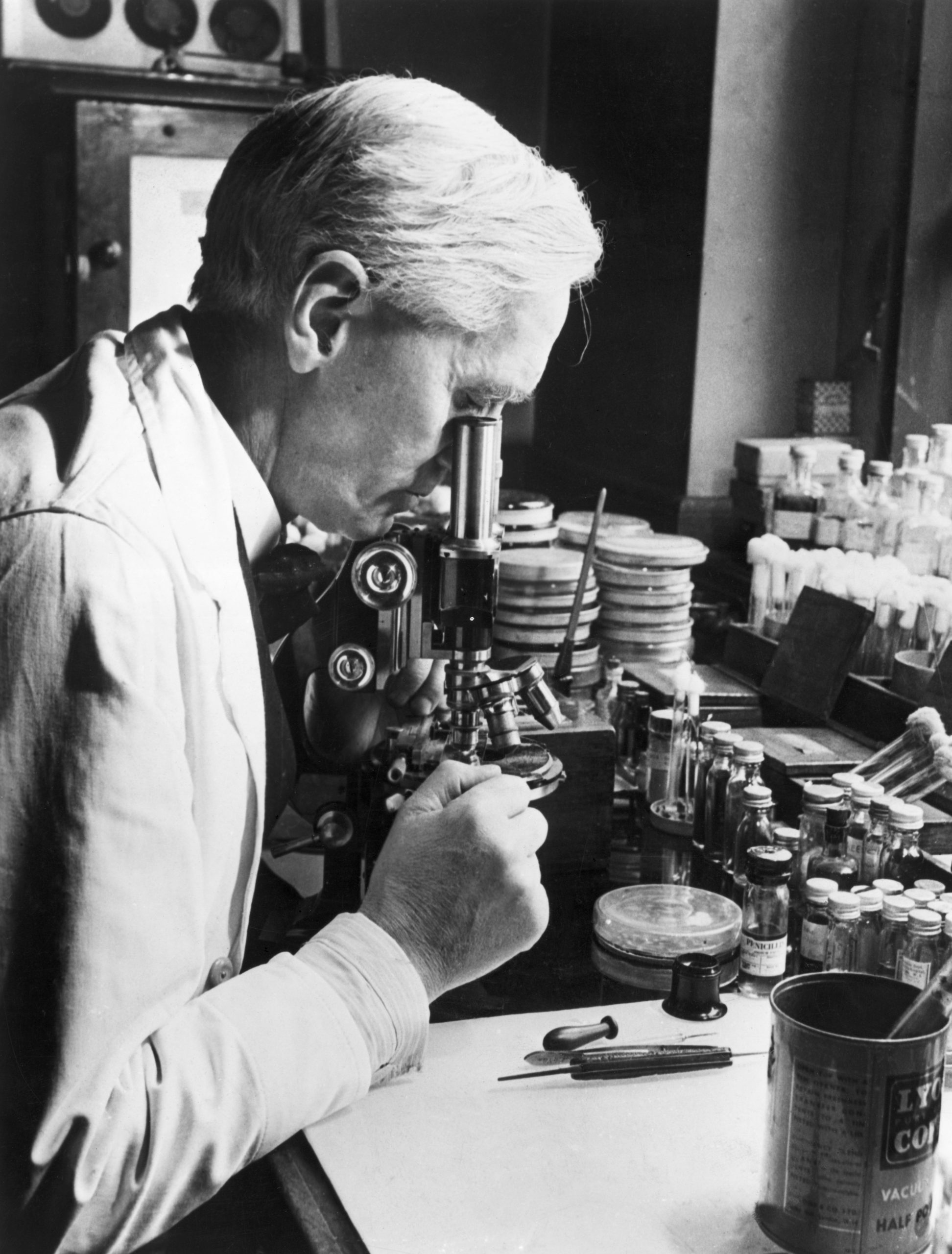 Foto do cientista Alexander Fleming dentro de um laboratório. Ele olha pela lente de um microscópio e ao seu redor há vários instrumentos, tubos de ensaio e placas.
