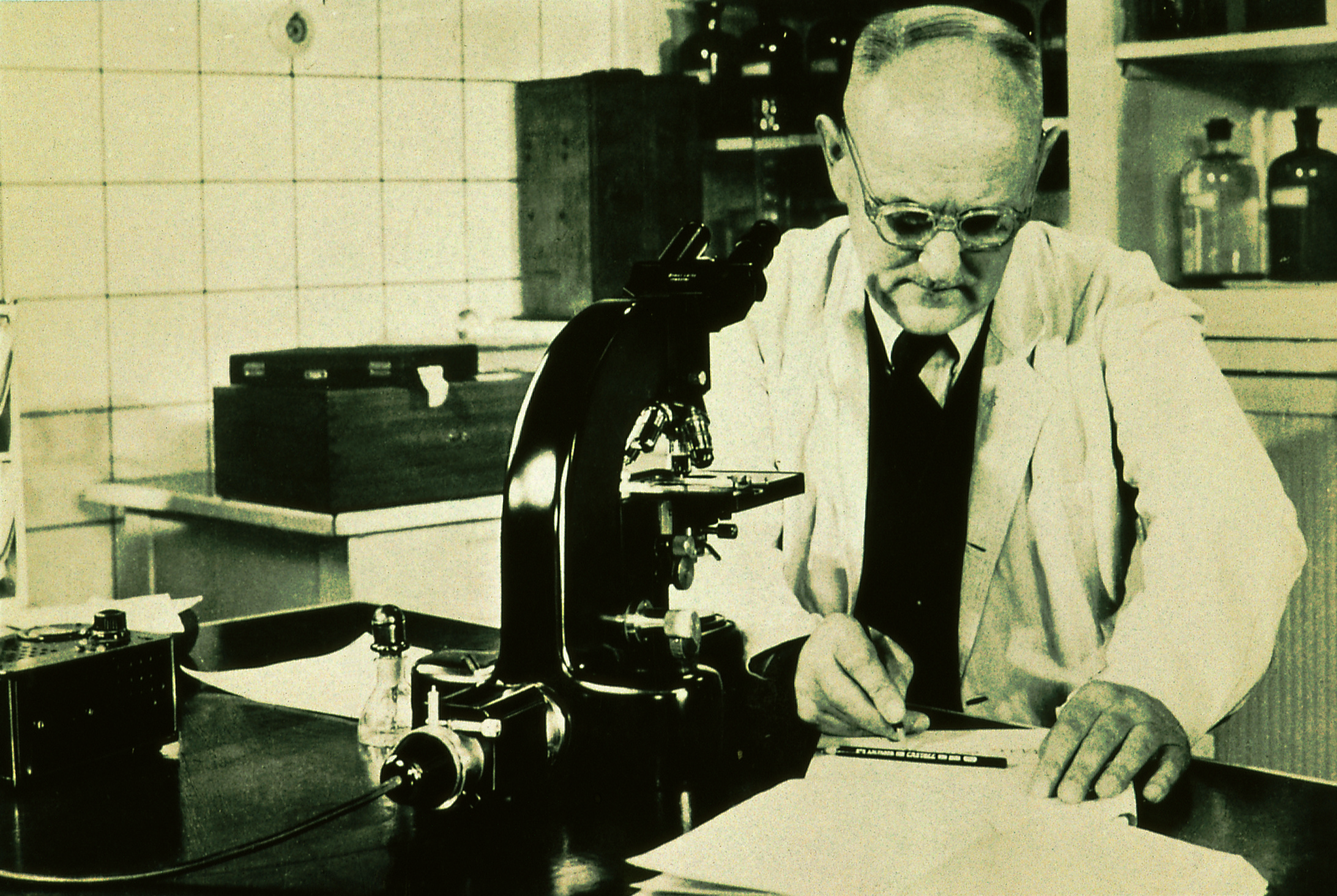 Foto em preto e branco do bacteriologista Gerhard Domagk dentro de um laboratório, escrevendo sobre uma folha, em cima de uma mesa onde há um microscópio. Ele está de óculos e veste um jaleco branco por cima de um colete e gravata.