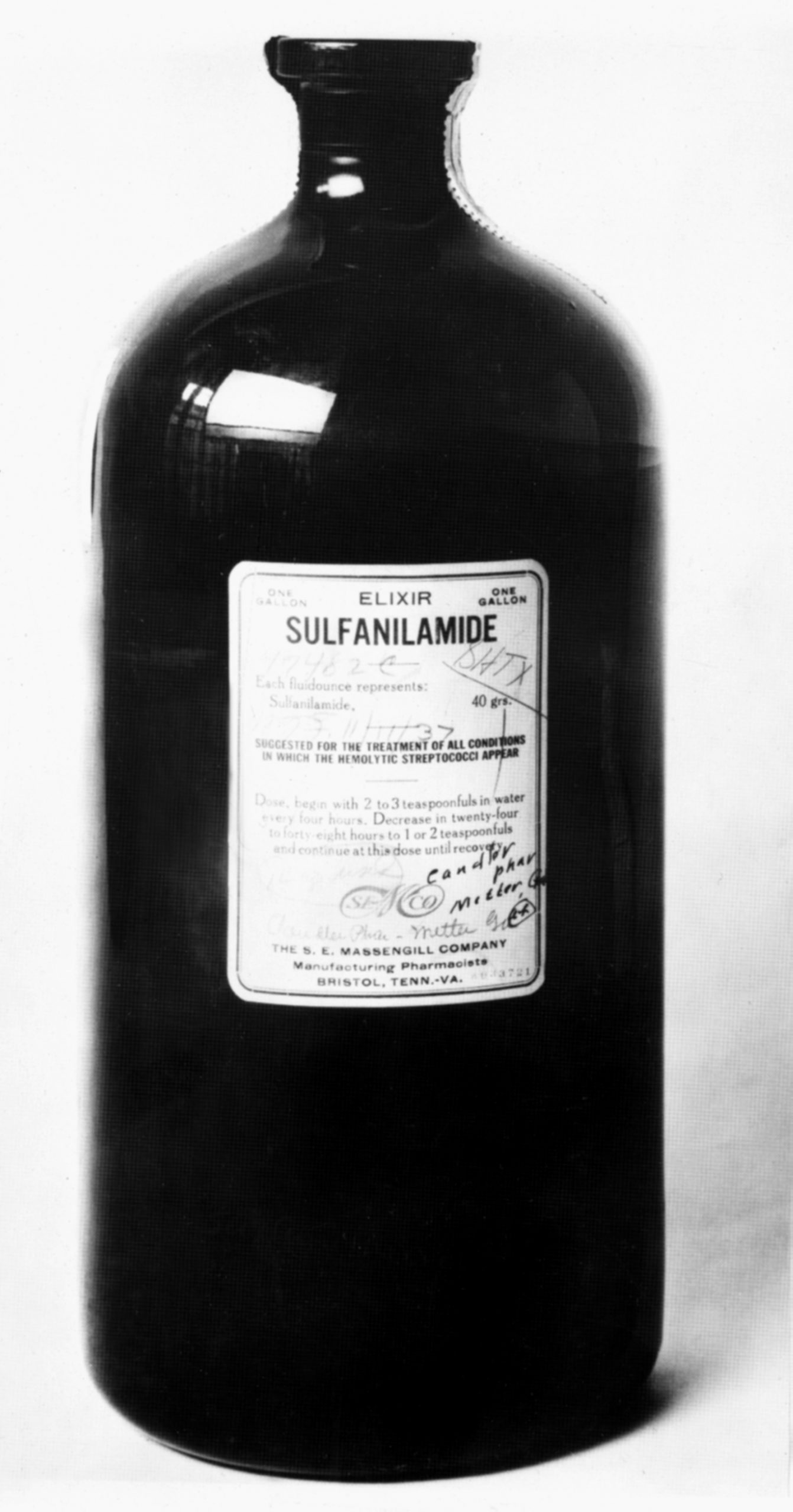 Foto em preto e branco de um frasco escuro, de vidro e com rótulo retangular do elixir Sulfanilamide.