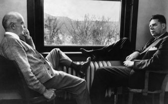 Foto em preto e branco de Edward Kendall e Phillip Hench sentados em cadeiras, um de frente para o outro, se olhando. Ambos colocam uma de suas pernas para cima, flexionadas, apoiadas em uma mobília próxima a uma janela.