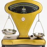Foto de uma balança em metal amarela, com um prato em cada lado, com um visor para indicar a pesagem  no centro.