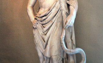 Estátua do deus Asclépio. Ele está de pé, usando uma túnica e ao seu lado há uma cobra com a cara próxima de sua mão.