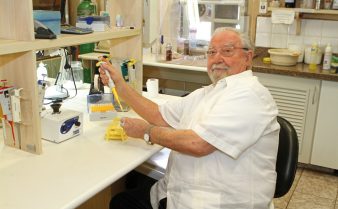 Foto do farmacologista Sérgio Ferreira, um homem branco, de cabelos, barba e bigode brancos, que usa um jaleco e óculos de grau, sentado em um laboratório. Ele segura uma pipeta e sorri para a foto.