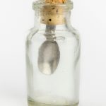 Foto de um frasco de vidro incolor, com boca estreita. Ele tem uma rolha com uma colher de metal acoplada nela.