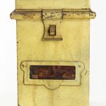 Foto de lata de metal antiga na cor amarela, quadrada e com a boca larga com marcas que lembram ferrugem e tinta descascada.