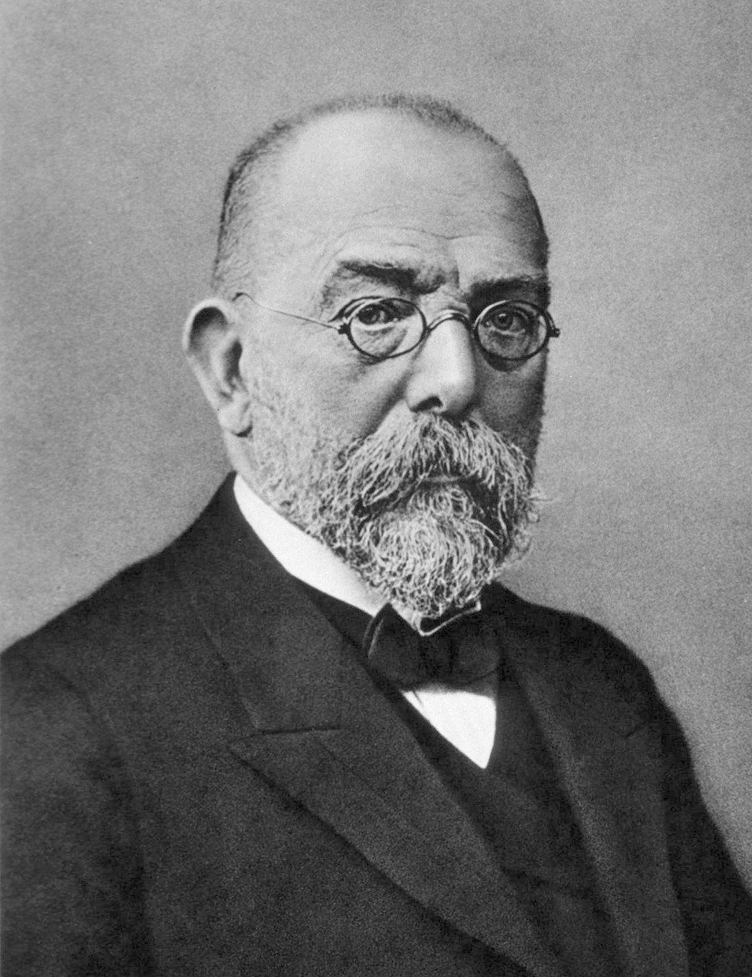 Foto antiga e em preto e branco do patologista alemão Robert Koch. Ele está de perfil, tem cabelos curtos, barba e bigode, usa óculos de grau com hastes arredondadas e veste roupa social.