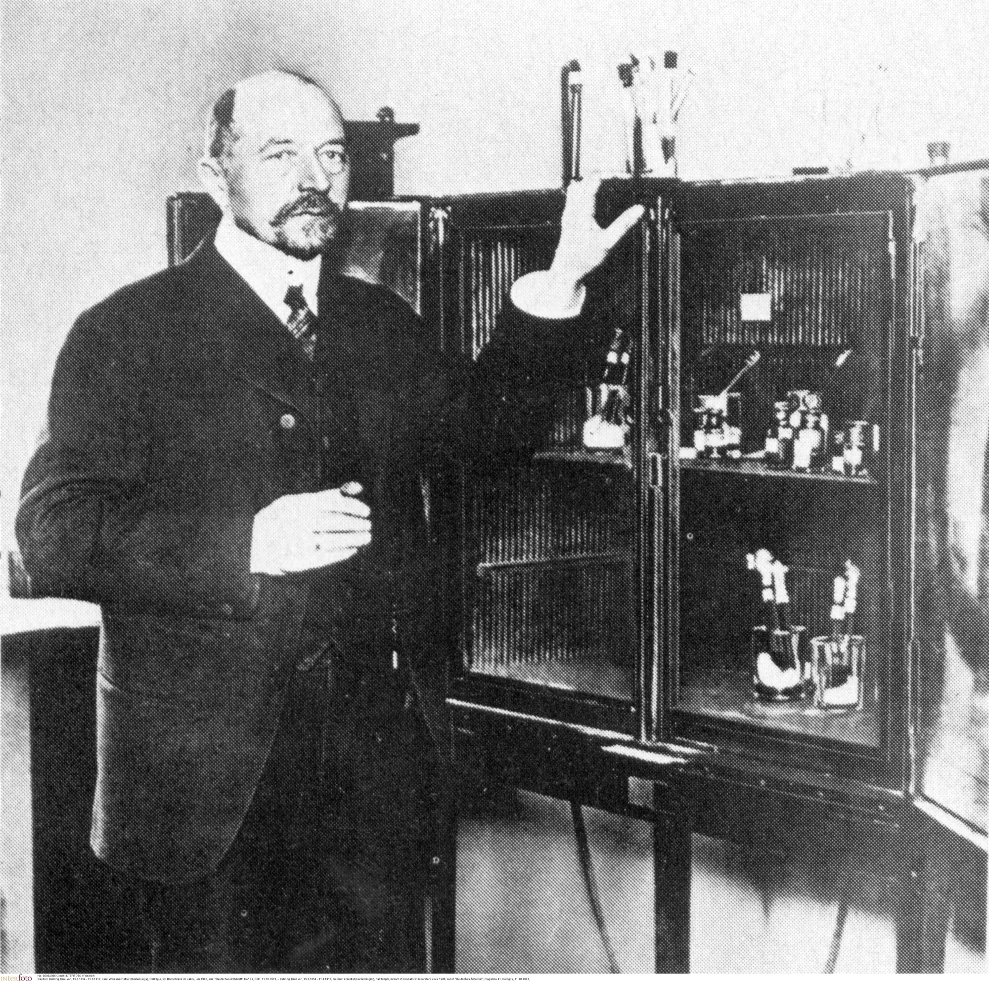 Foto em preto e branco do médico alemão Emil von Behring ao lado de uma estante de altura média, cheia de medicamentos. Emil é calvo, usa barba e bigode, e veste terno e gravata.