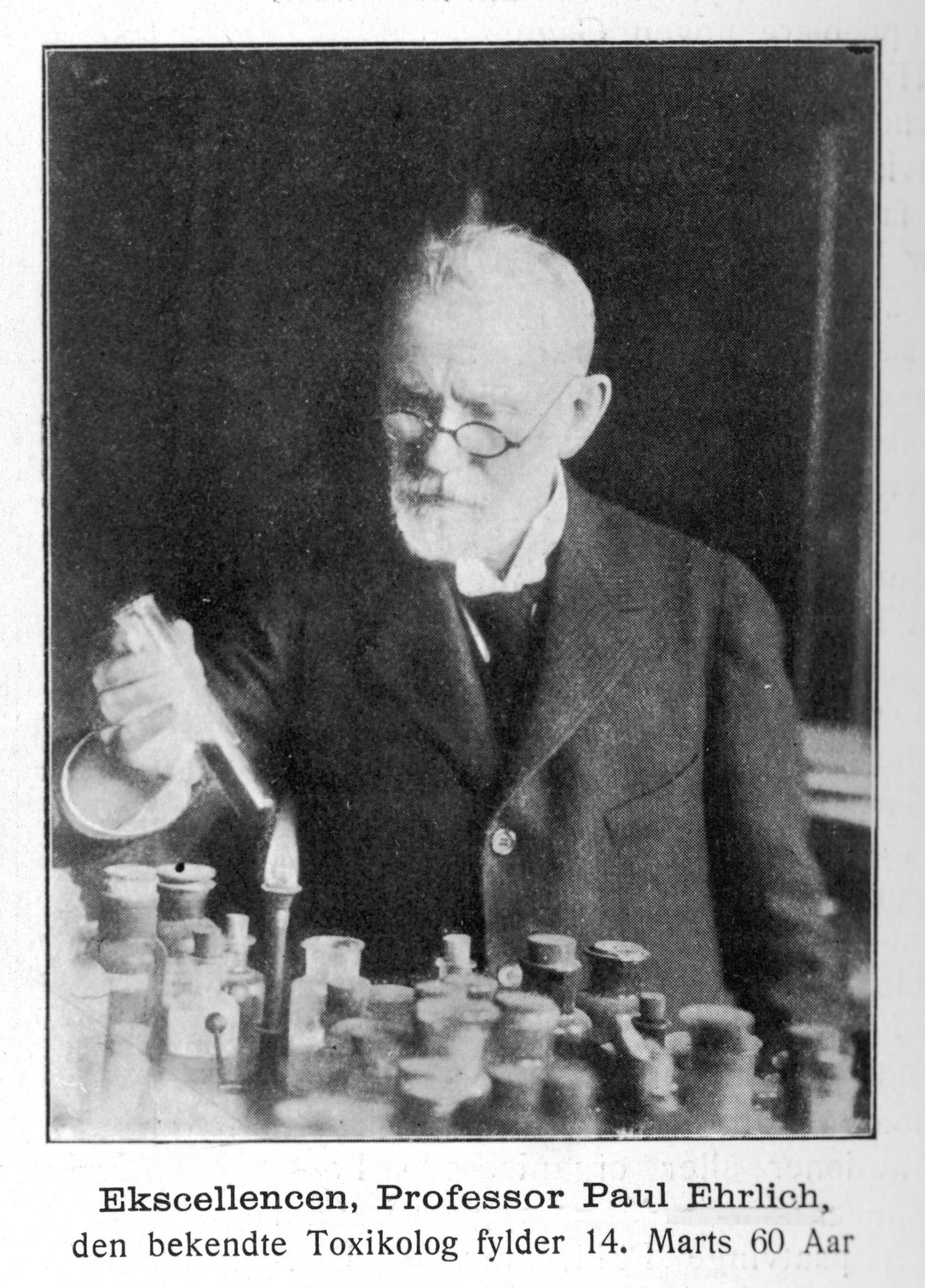 Foto em preto e branco do fisiologista Paul Ehrlich em frente a diversos frascos sobre uma mesa. Ele tem uma pipeta na mão, usa terno e gravata e óculos de grau.