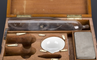 Foto de um kit de Salvarsan para tratamento da sífilis, com uma garrafa medidora de vidro, uma porcelana branca côncava, uma lata retangular e um conta gotas.