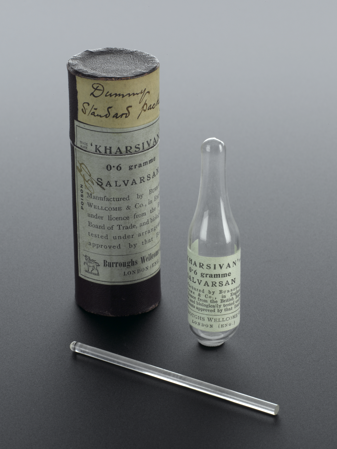 Foto de uma garrafa de vidro de Salvarsan com rótulo, ao lado da embalagem do produto em formato cilíndrico, e um bastão de vidro com ponta polida.