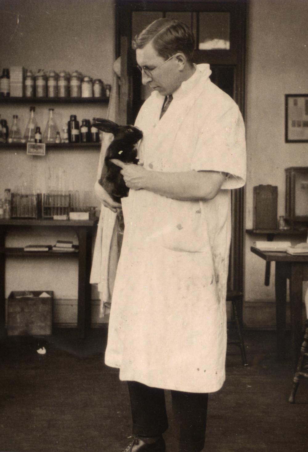Foto em preto e branco de Frederick Grant Banting dentro de um laboratório, de jaleco, segurando um coelho preto.