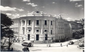 Foto em preto e branco da fachada da Faculdade de Medicina, em Porto Alegre, no Rio Grande do Sul, em registro anterior a 1965. O prédio fica em uma esquina e ocupa uma quadra. Ao seu redor há árvores e alguns carros estacionados, além de pessoas andando na rua e na calçada.