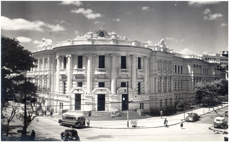 Foto em preto e branco da fachada da Faculdade de Medicina, em Porto Alegre, no Rio Grande do Sul, em registro anterior a 1965. O prédio fica em uma esquina e ocupa uma quadra. Ao seu redor há árvores e alguns carros estacionados, além de pessoas andando na rua e na calçada.