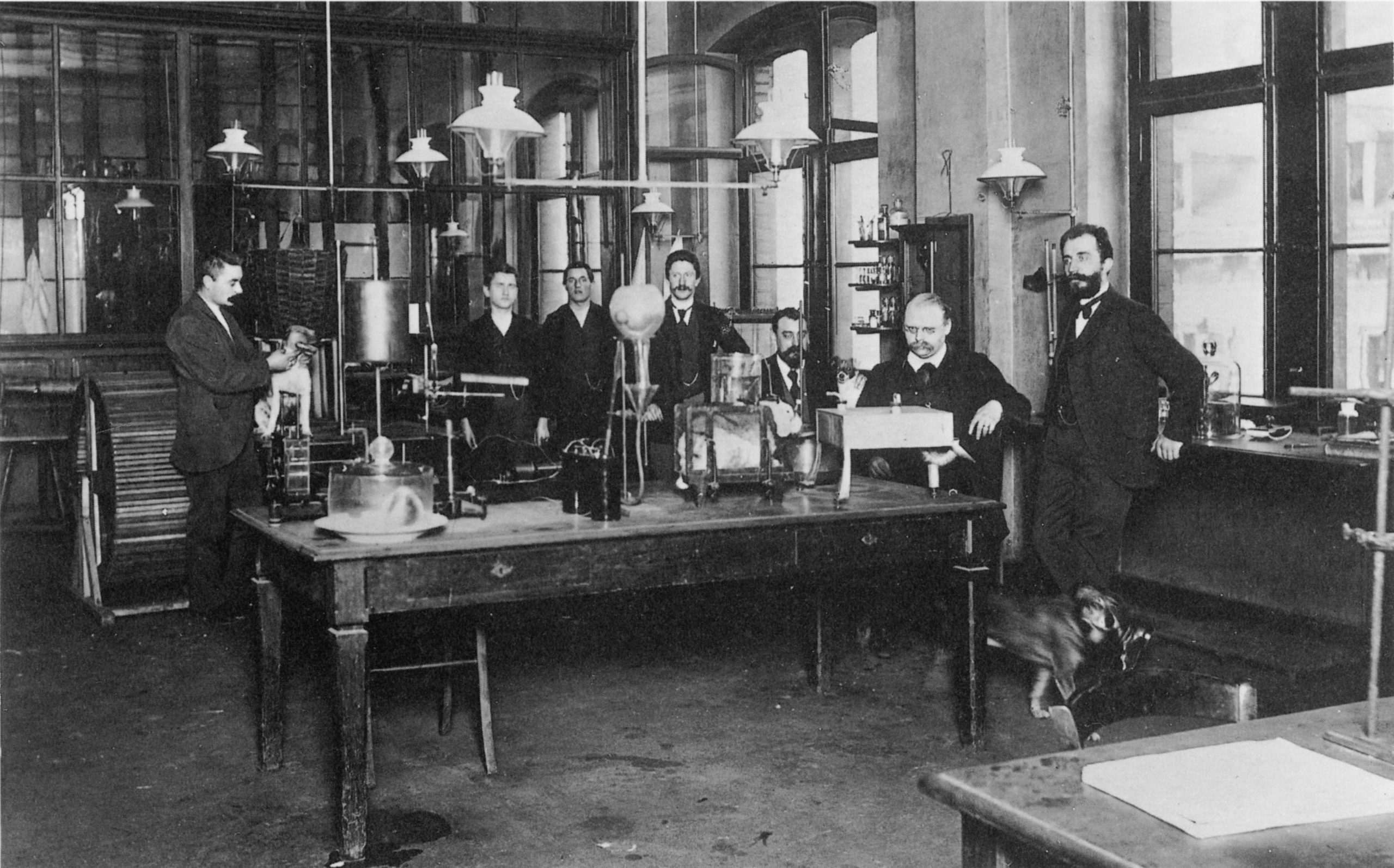 Foto em preto e branco do interior do laboratório da Bayer. Sobre a mesa central, há vários objetos de uso laboratorial, e ao redor dela, sete homens vestidos de terno e gratava.