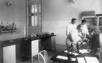Foto do bacteriologista Oswaldo Cruz trabalhando com duas outras pessoas em um laboratório. Há mesas, armários e prateleiras com frascos, tubos e outros objetos.