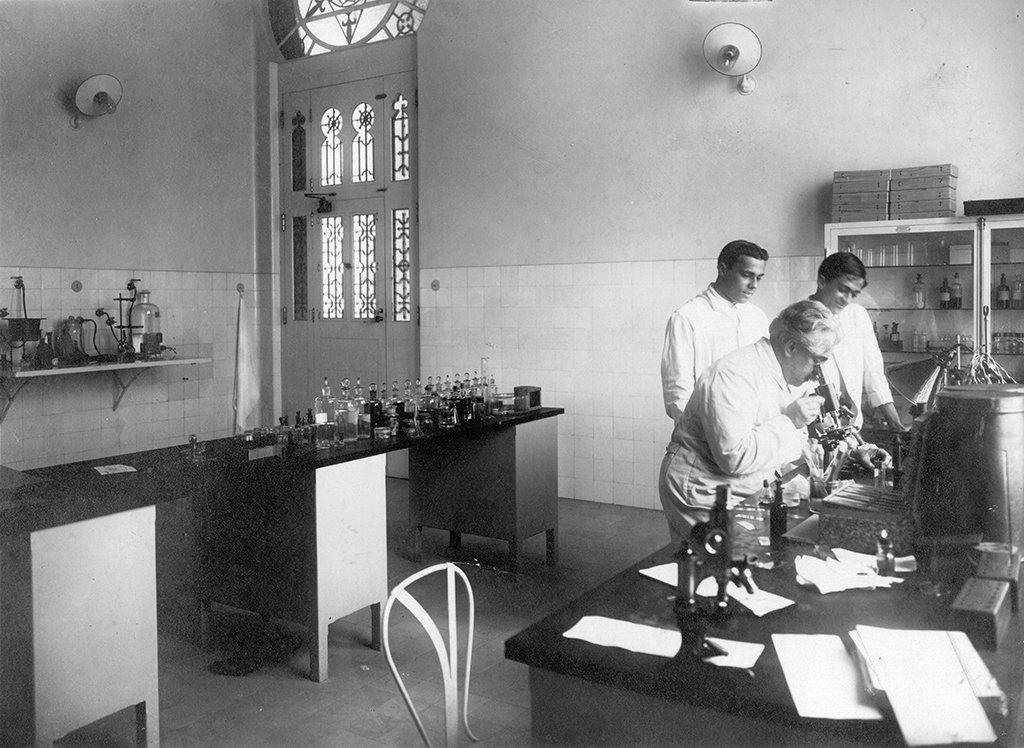 Foto do bacteriologista Oswaldo Cruz trabalhando com duas outras pessoas em um laboratório. Há mesas, armários e prateleiras com frascos, tubos e outros objetos.