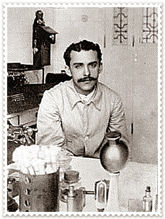 Foto em preto e branco do médico Gaspar de Oliveira Vianna. Ele está num laboratório sentado em frente à uma mesa com objetos variados, e  usa camisa, tem os cabelos curtos, ondulados e penteados para trás e longos bigodes pretos.