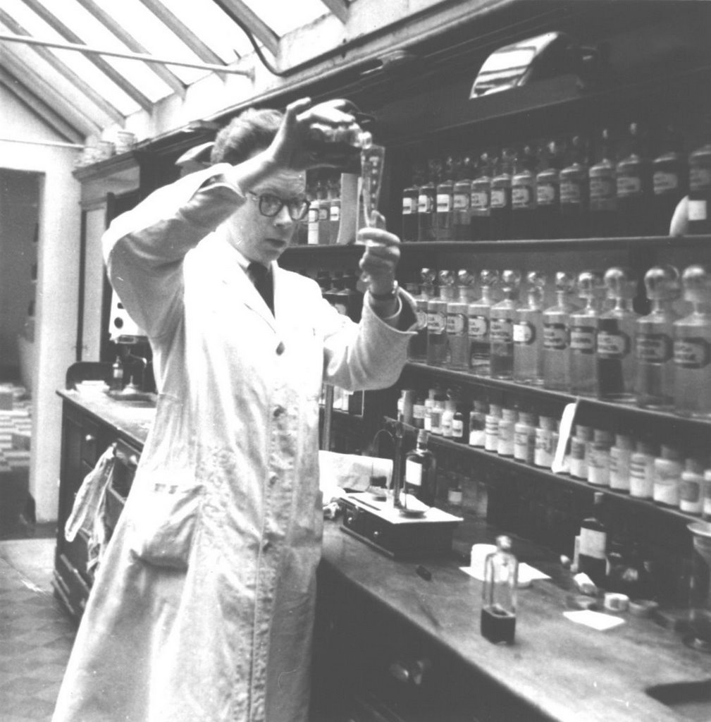 Foto em preto e branco do interior de um laboratório com um farmacêutico, de jaleco, colocando um produto de um vidro pequeno dentro de um tubo de ensaio. Ao lado dele, uma prateleira grande cheia de garrafas de vidro e potes de medicamentos.