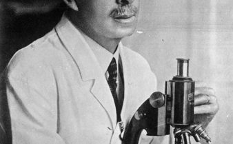 Foto em preto e branco do médico Carlos Chagas dentro de um laboratório, olhando para a frente enquanto tem as mãos em um microscópio. Ele está de jaleco branco, tem cabelos curtos e penteados para trás, e usa bigode.