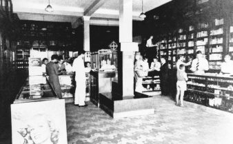 Foto em preto e branco do interior de uma farmácia, com balcões, colunas, prateleiras, atendentes e consumidores.