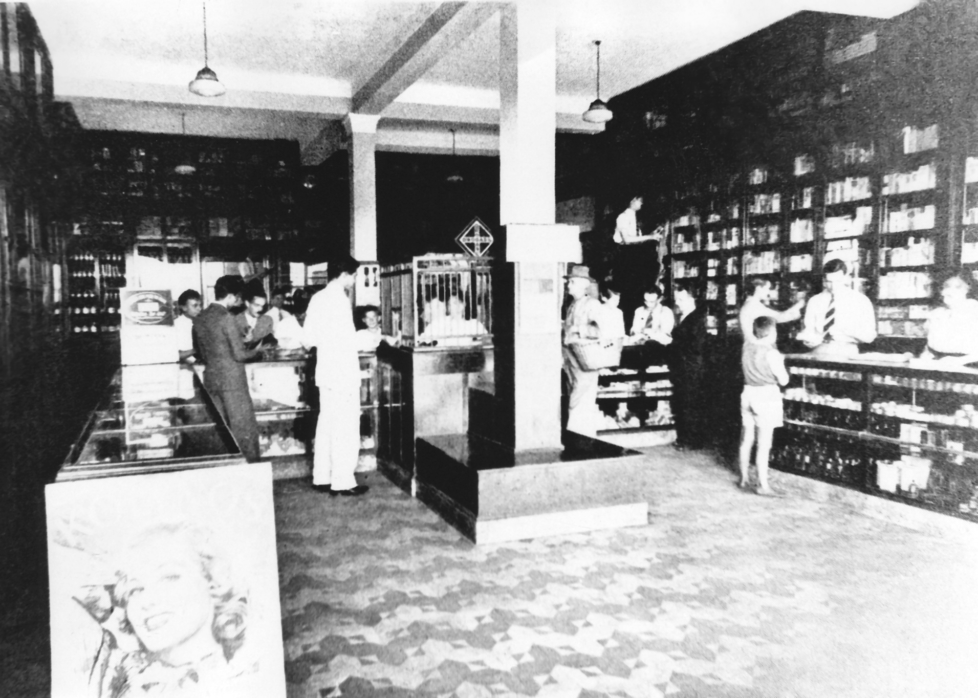 Foto em preto e branco do interior de uma farmácia, com balcões, colunas, prateleiras, atendentes e consumidores.