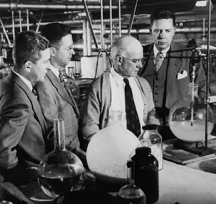 Foto em preto e branco do bioquímico norte-americano Edward Calvin Kendall em um laboratório com outros três homens. Todos vestem terno, gravata e colete, e olham em direção a um balão de destilação de vidro com uma composição química dentro. Ao lado, há, sobre a mesma mesa, outros balões de vidro com substâncias.