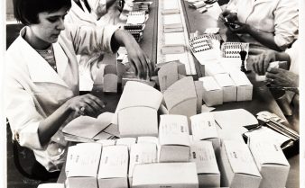 Foto em preto e branco da linha de produção de pílulas anticoncepcionais na Inglaterra, em 1955. Há muitas mulheres concentradas, de cabeças levemente baixas, vestindo o mesmo tipo de casaco de cor clara, ao redor de uma mesa longa, repleta de caixas.