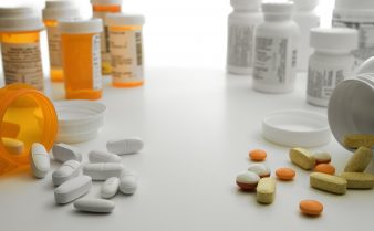 Foto de frascos de remédios nas cores laranja e branco, alguns abertos, com comprimidos, brancos e coloridos, espalhados numa mesa branca.