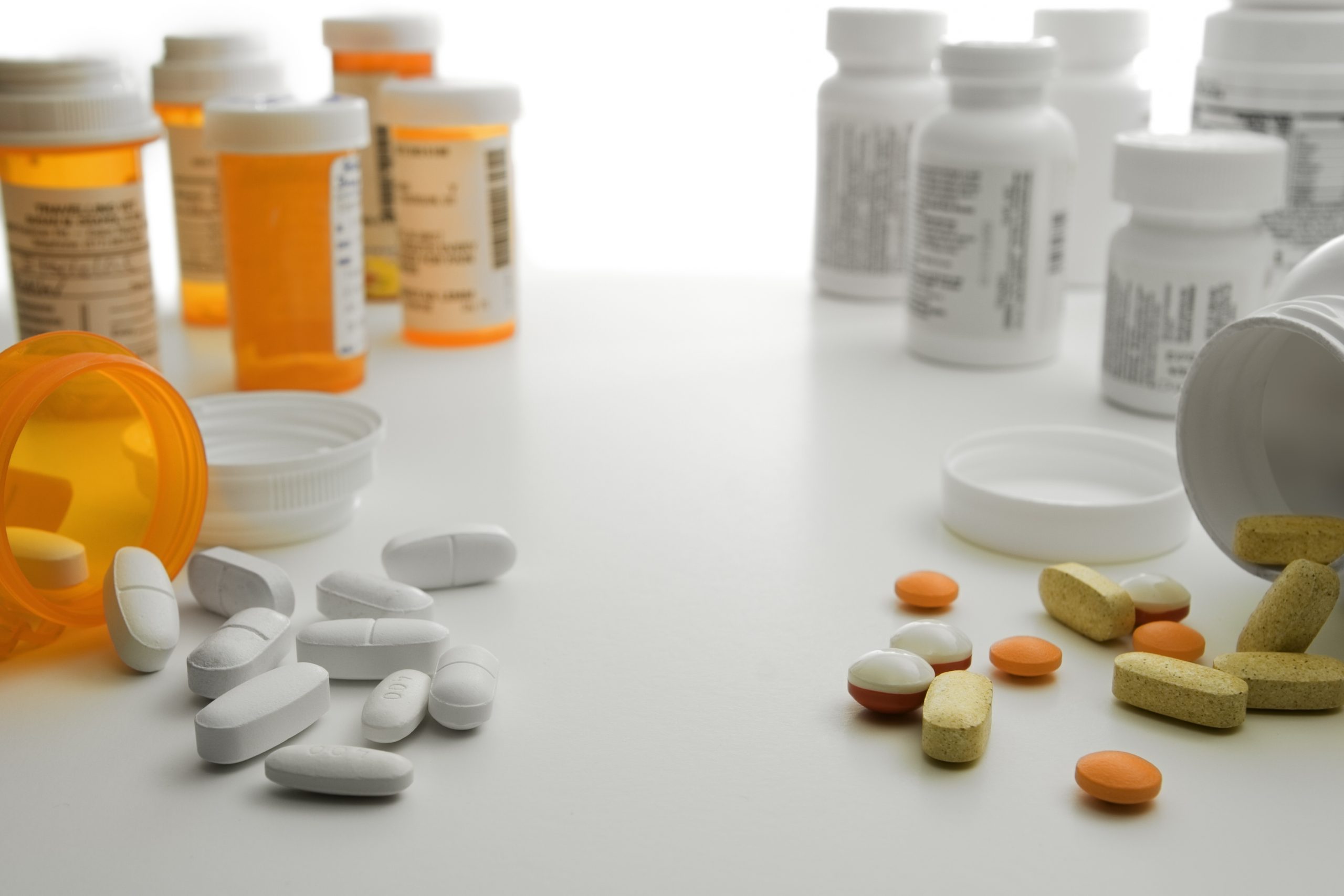 Foto de frascos de remédios nas cores laranja e branco, alguns abertos, com comprimidos, brancos e coloridos, espalhados numa mesa branca.