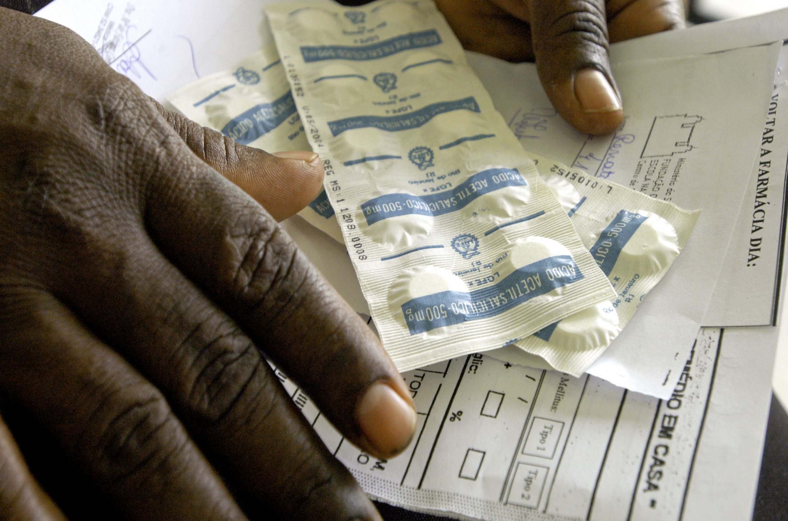 Foto com as mãos, em close, de uma pessoa negra segurando duas cartelas de comprimidos e papeis de receitas médicas.