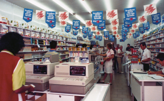 Foto do interior de uma loja da Droga Raia nos anos 1980 com caixas registradoras, clientes ao redor das gôndolas e bandeirinhas publicitárias penduradas no teto.
