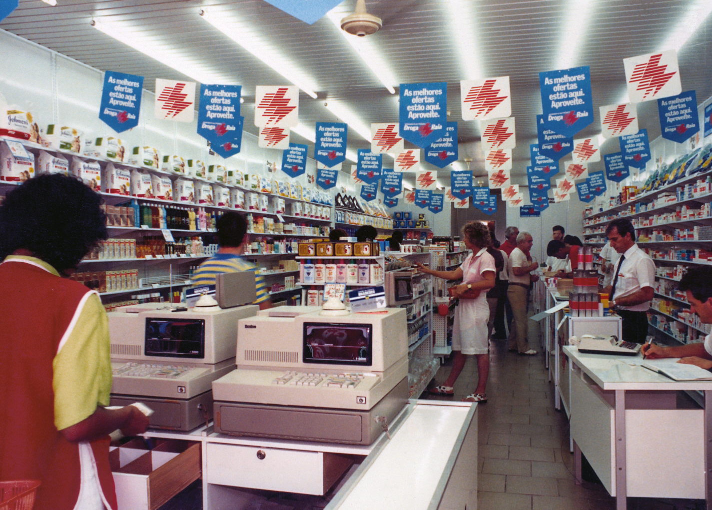 Foto do interior de uma loja da Droga Raia nos anos 1980 com caixas registradoras, clientes ao redor das gôndolas e bandeirinhas publicitárias penduradas no teto.