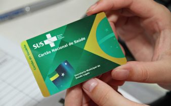 Foto de mãos segurando um cartão Nacional de Saúde do SUS. O cartão é verde, tem o logo do SUS no canto superior esquerdo e a metade esquerda da bandeira do Brasil no canto direito.