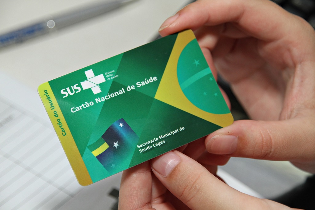Foto de mãos segurando um cartão Nacional de Saúde do SUS. O cartão é verde, tem o logo do SUS no canto superior esquerdo e a metade esquerda da bandeira do Brasil no canto direito.