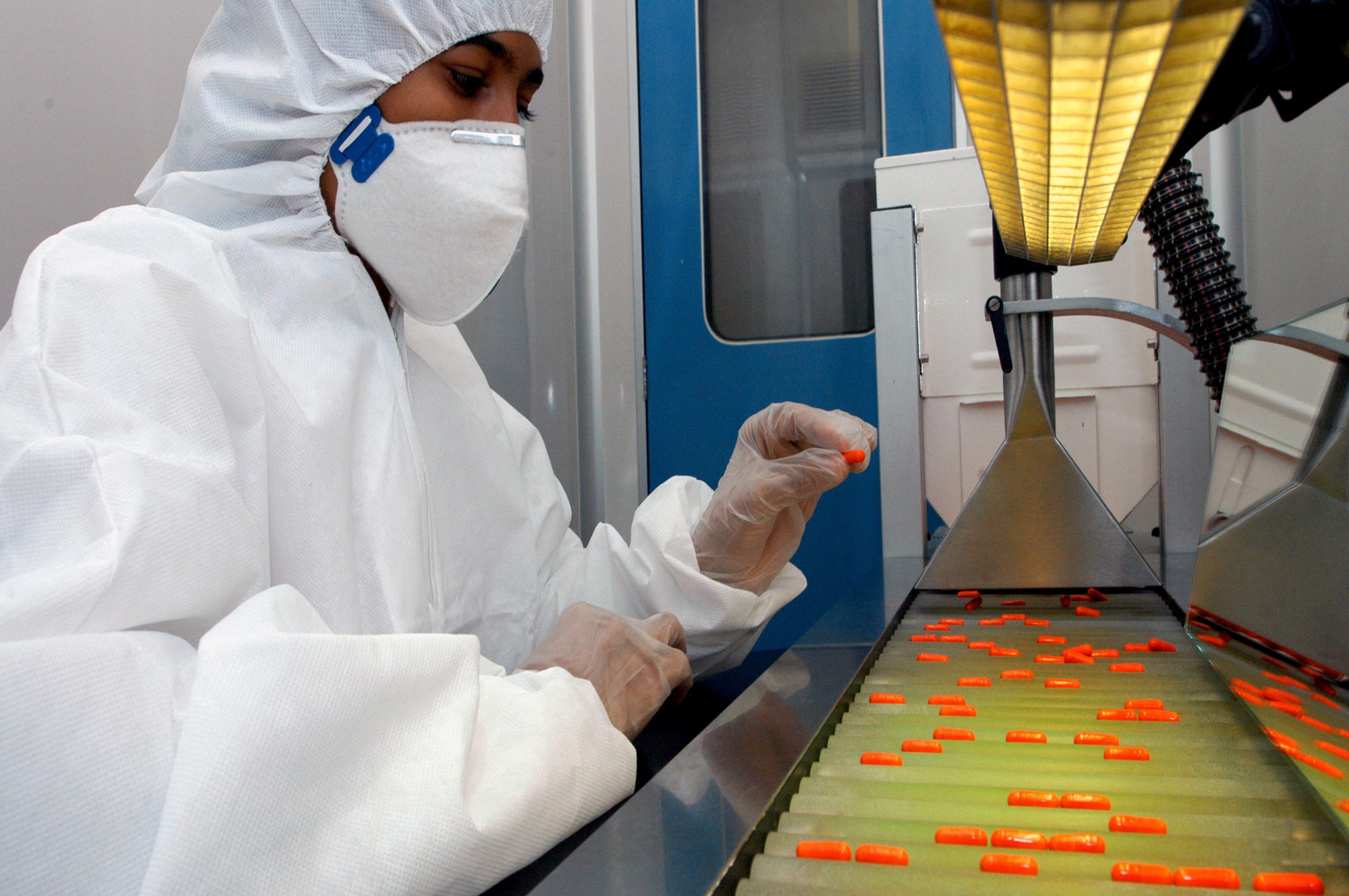 Foto de um pesquisador em um laboratório. Ele usa macacão, luvas  e máscara brancos, para proteção, e está em frente à uma máquina com uma mini esteira cheia de medicamentos cor de laranja.