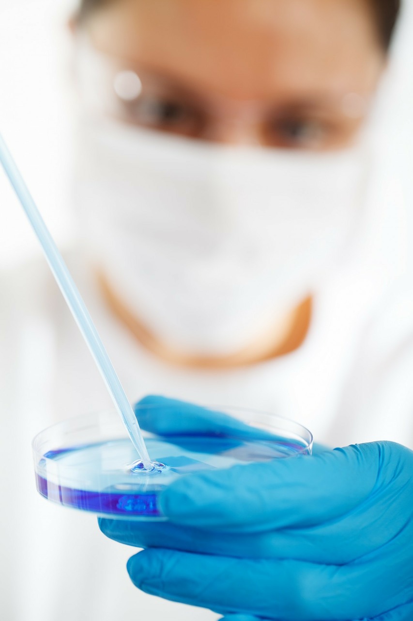 Foto de um pesquisador, em segundo plano, dentro de um laboratório, usando máscara  e óculos de proteção, segurando uma placa de Petri e uma pipeta de vidro. A mão, com luvas de látex azuis, segurando a placa, aparece em primeiro plano.