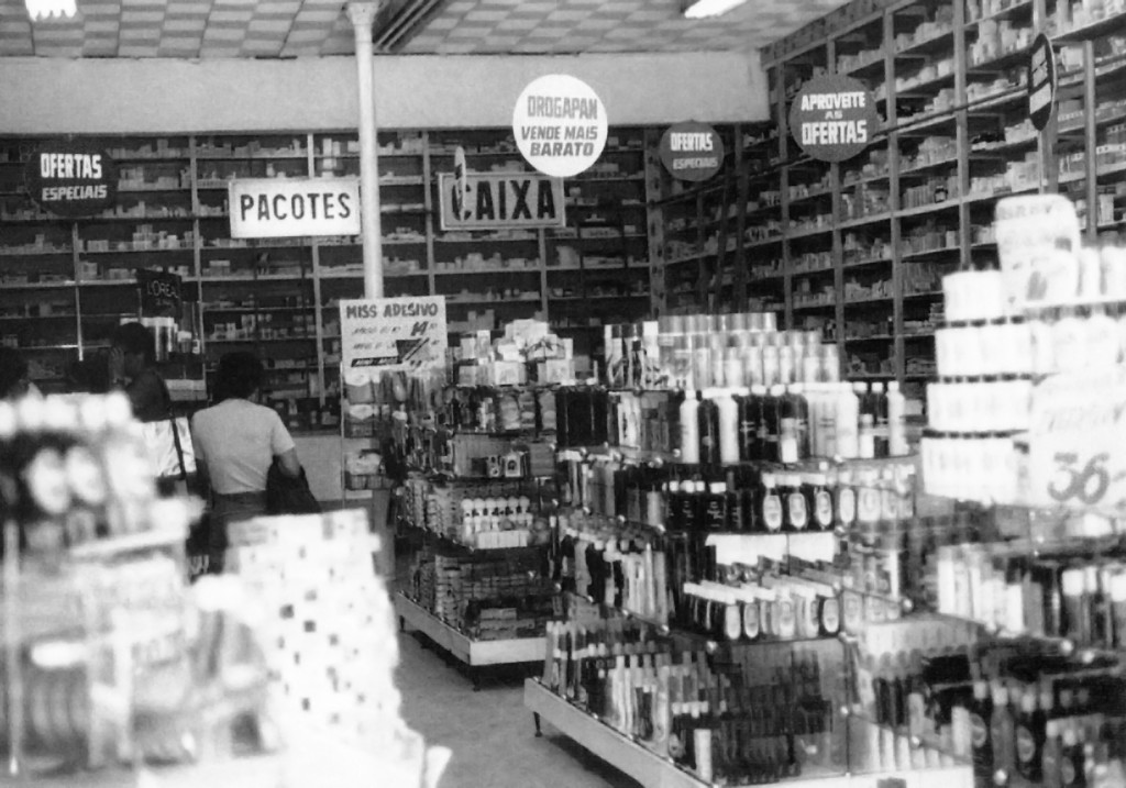 Foto antiga, em preto e branco, de uma drogaria. Há gôndolas, estantes e um balcão ao fundo, todos repletos de produtos. Ao fundo, há uma placa suspensa onde se lê "Pacotes".